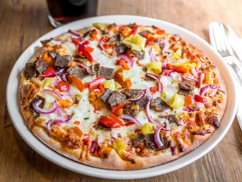 A pizza topped with sliced beef, red onions, red and yellow peppers, and melted cheese on a white plate, with utensils and a drink in the background.