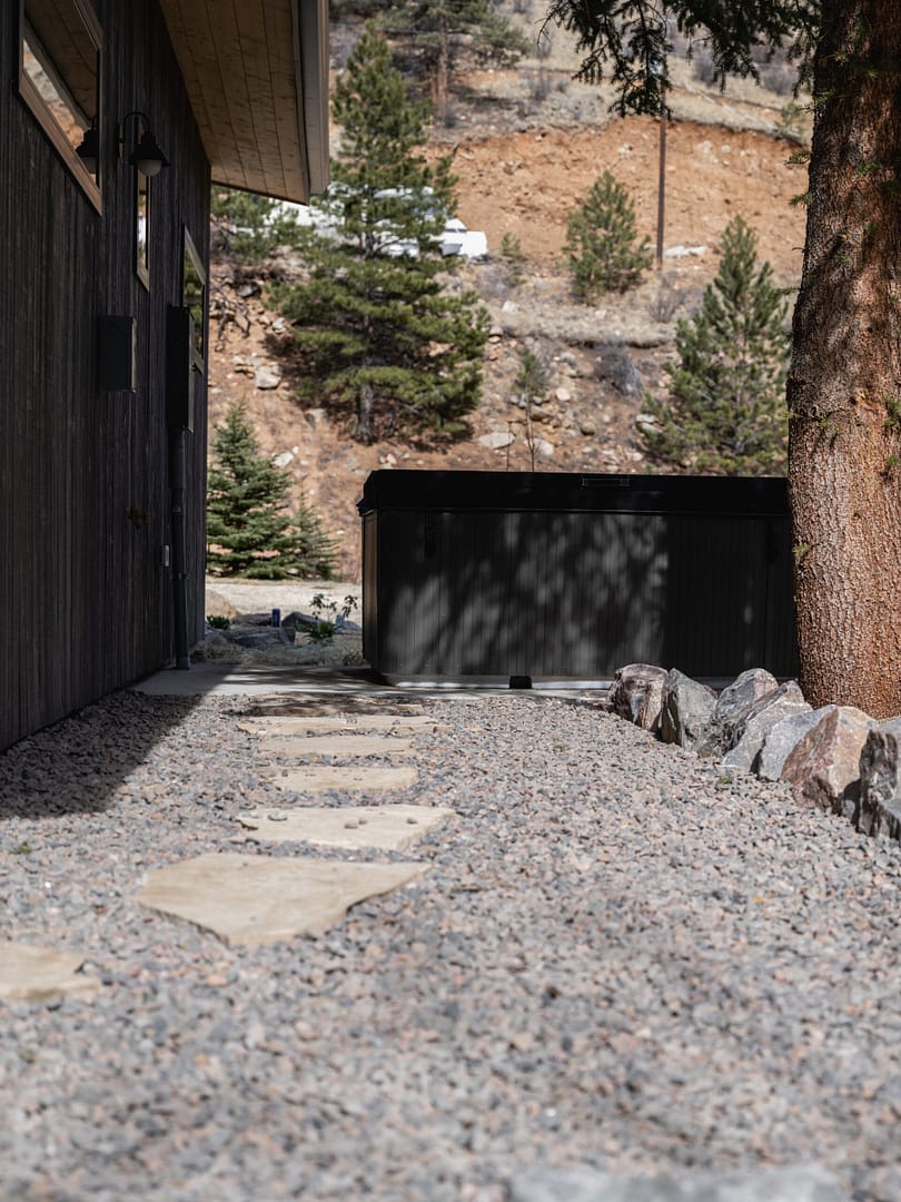 A stone path leads alongside a dark wooden building, bordered by rocks and gravel, toward a black hot tub. Tall pine trees and a rocky, sloped landscape are visible in the background.