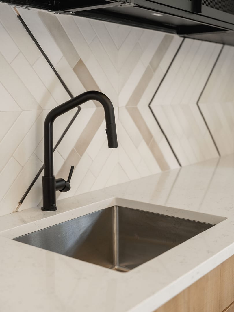 A modern kitchen sink with a matte black faucet set in a white countertop, featuring geometric, diagonal-patterned beige and white backsplash tiles.