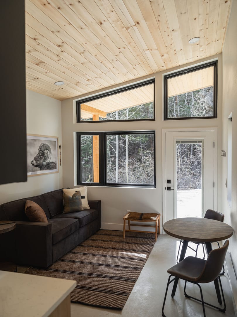 A cozy living area with a black sofa, striped rug, small round dining table with two chairs, large windows, and a glass door showing a snowy forest outside. The ceiling is light wood, adding warmth to the space.