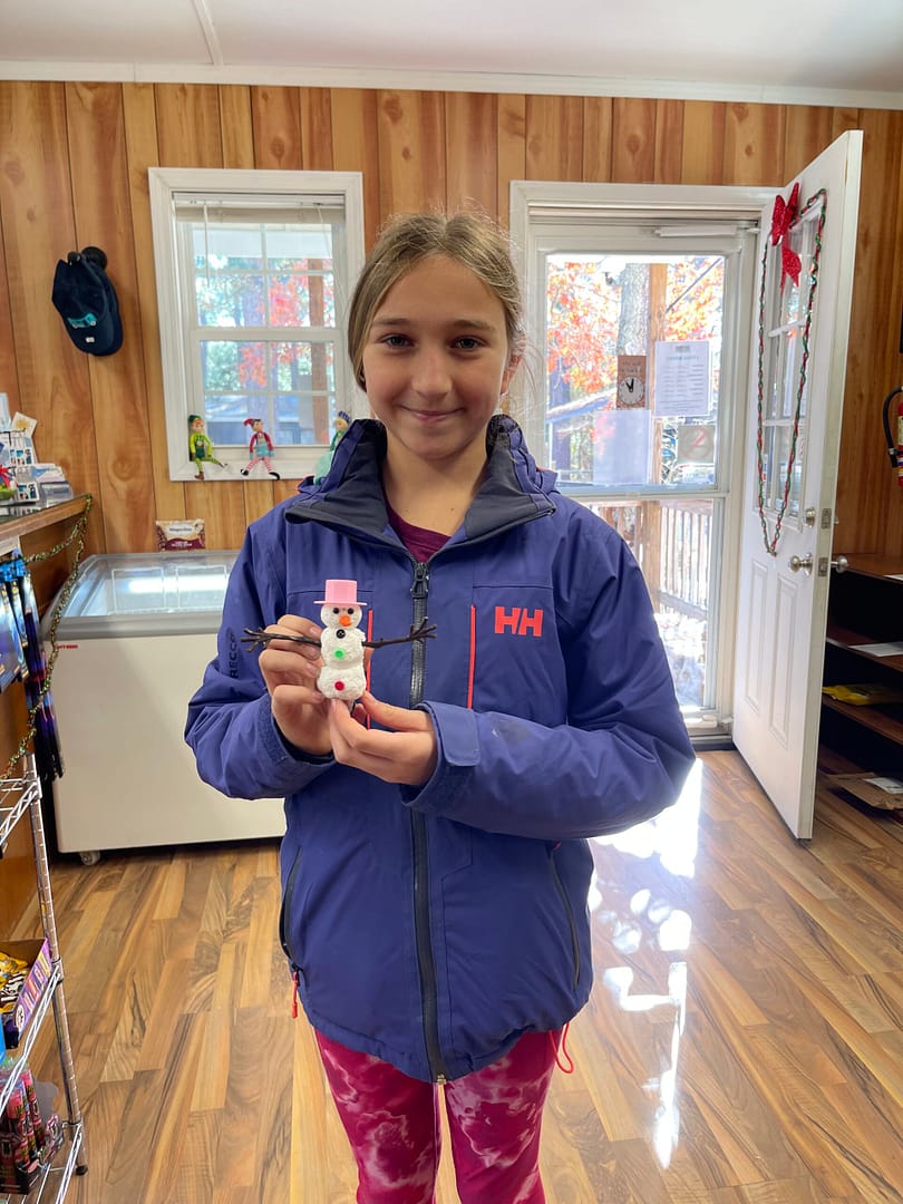 A young girl in a blue jacket holds a small snowman figurine inside a cozy craft center with wood-paneled walls and a glass door. She smiles while standing on the wooden floor, where shelves and various items surround her, creating an inviting atmosphere for cosmic kids.