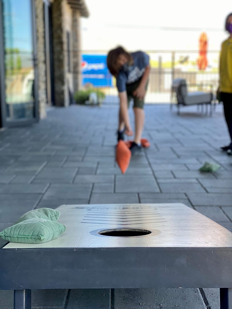 A person throws an orange bean bag toward a cornhole board outdoors, with two green bean bags resting on the board in the foreground.