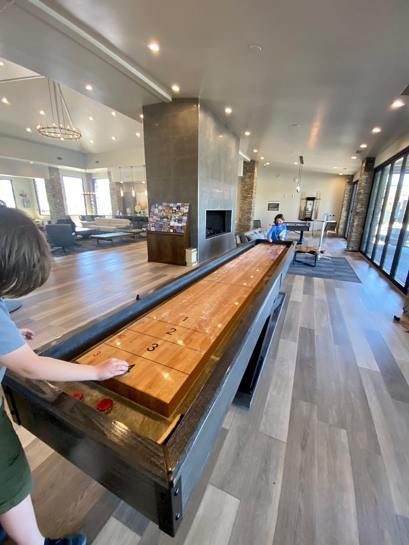 Two people play shuffleboard on a wooden table in a modern, spacious room with large windows and wood flooring.