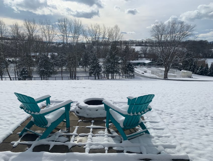 Two turquoise Adirondack chairs face a snow-covered fire pit on a patio, overlooking a wintry landscape with bare trees and cloudy sky.