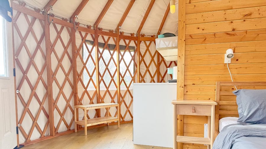 Interior of a bright yurt with wooden walls, flooring, and furniture, featuring a bed and natural light.