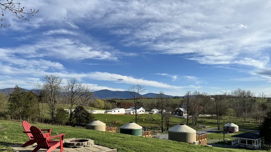 Rural landscape with yurts, a fire pit, and red chairs overlooking mountains.