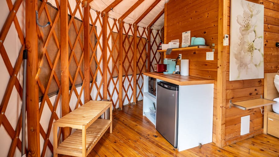 A small, cozy yurt interior with wood lattice walls, a mini fridge, microwave, countertop appliances, shelves with dishes, a wooden bench, and a painting of white flowers on the wall.