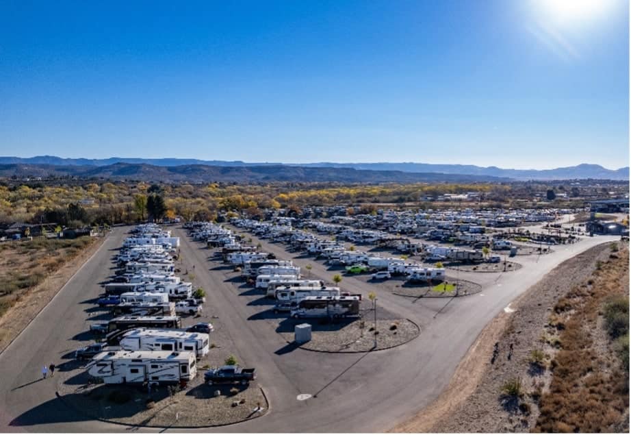 Aerial view of a large RV park with multiple rows of parked recreational vehicles under a clear blue sky, surrounded by natural landscape.