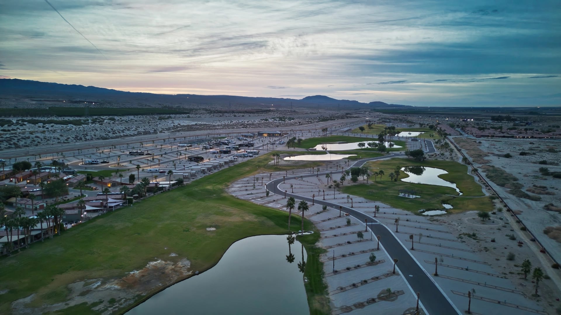 Aerial view of an empty parking lot, green spaces, small ponds, and scattered palm trees under a cloudy sky at dusk.